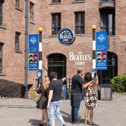 Outside of The Beatles Story with people stood by the entrance. There are blue banners either side of the staircase leading down into the attraction.