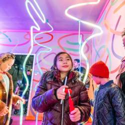 Inside a colourful pink box. There are ribbon shaped LED lights hanging from the ceiling. Children are smiling looking at the lights and an adult is watching the children smiling.