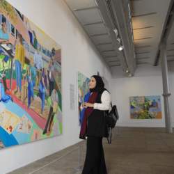 A lady wearing long black clothes looks at a large colourful and lifelike painting in a gallery. The gallery has white walls and grey metal supports.