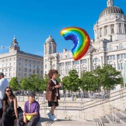 A family on the Liverpool Pier Head on a sunny day. A boy holding a rainbow balloon in front of the three graces buildings.