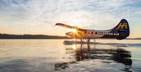 A seaplane taxiing on calm water at sunrise with sunlight shining through its wing.