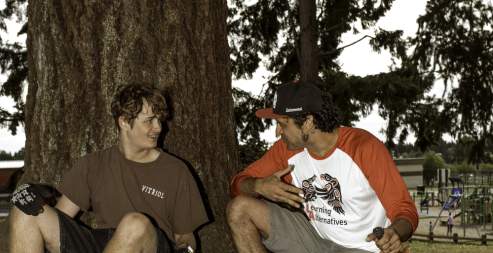 Two men sitting leaning on a tree with a bike.