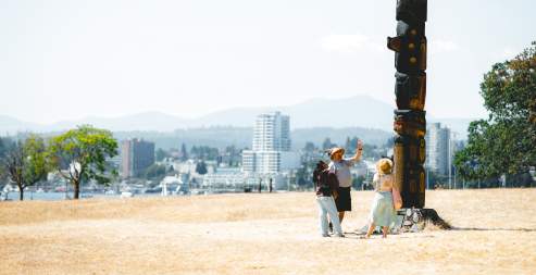 Three people stand at the base of a totem pole on Saysutshun with Nanaimo’s skyline visible across the harbour in the background.