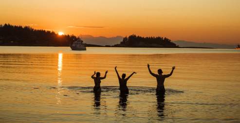 Three people standing in calm ocean water at sunset with arms raised.