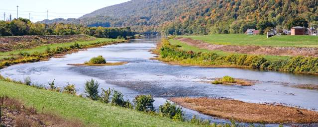 River winding through a grassy valley with tree-covered mountains in the background under a clear blue sky.