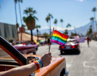 Pride flag waving outside of car in downtown Palm Springs with mountains and palm tree