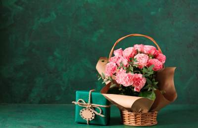 A wicker basket filled with pretty pink flowers next to a small square parcel wrapped in green paper and string with a gift tag