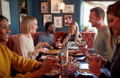 A group of five friends cheerfully gathered around a table in a cosy restaurant, enjoying a meal. Warm lighting and framed art create a welcoming atmosphere.