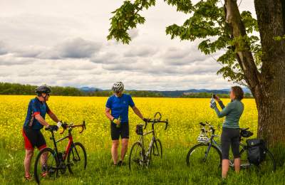 Three cyclists stand beside their road bikes in front of a field of bright yellow flowers under a tree at Camas Country Mill.