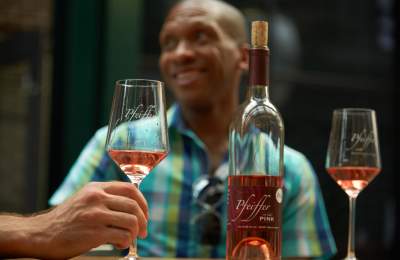 In the foreground there is a bottle and two glasses of rosé. The bottle says Pfeiffer. In the background and out of focus is a man smiling.