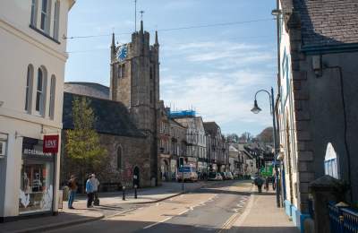 A quaint street scene features a stone church with a tall clock tower, flanked by shops and buildings; people walk under a bright blue sky.