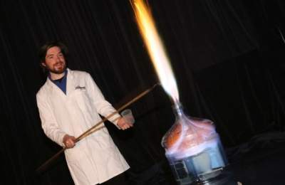 Man in a white labcoat holds a long wooden stick over an open flame coming out of a water cooler.