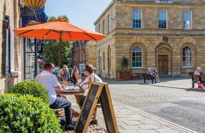 A group of people sit in the shade of an orange parasol outside a cafe, people sit on benches outside the adjacent town hall, whilst two ladies walk along shopping. Woodstock in the Cotswolds