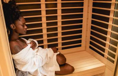 A black woman sits peacefully in a cedar sauna, wrapped in a white robe with eyes closed, enjoying a calming self-care moment in Fayetteville,