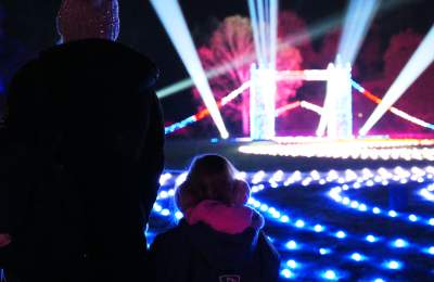 A mother and small child stand with their backs to the camera looking at a small, lit up London Bridge. There are roaming spotlights & glowing fairylights on the floor in all different colours. The trees off in the distance are also lit up in blue and pink.