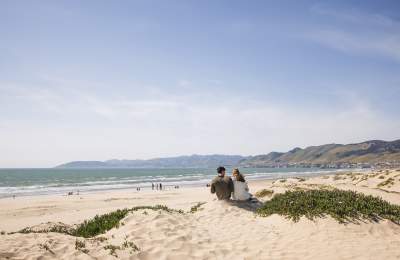 Two people sitting on the beach in Grover Beach with the coastline in the background