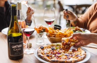 Table with wine bottles, glasses, and food. Person picking up a slice of pizza.