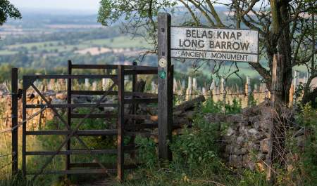 Kissing gate with footpath sign to Belas Knap with a stone wall ahead and behind a view of the valley