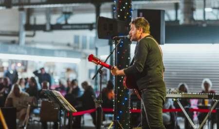 A singer stands on stage with a guitar in Trinity Market, Hull. People sit at tables in the background.