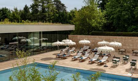 An outdoor swimming pool with rows of sunbeds and sun umbrellas, surrounded by trees at Cowley Manor, in the Cotswolds