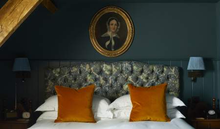 A teal coloured wall behind a double bed with burnt orange decorative cushions