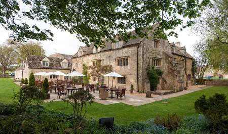 Wooden tables and chairs with white parasols sit on a patio behind The Slaughters Country Inn, surrounded by gardens
