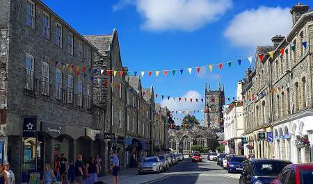 A vibrant street scene with colorful pennant flags strung between gray stone buildings under a bright blue sky. People and parked cars line the street.