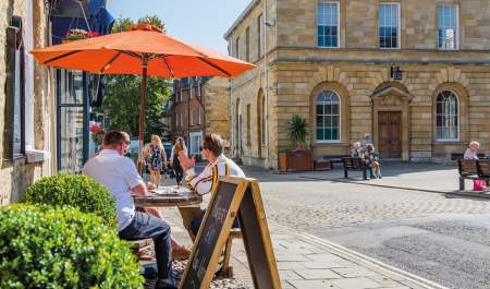 A group of people sit in the shade of an orange parasol outside a cafe, people sit on benches outside the adjacent town hall, whilst two ladies walk along shopping. Woodstock in the Cotswolds