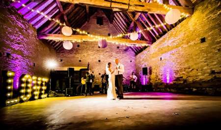 A newly married couple dance in the centre of a historic barn with fairy lights hanging from the wooden beamed ceiling at Cogges Manor Farm in the Cotswolds