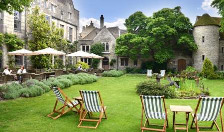 Traditional deckchairs sit on a lawn with flower borders. People sit drinking under sunshades on a patio in front of the 16th century town house at The Close Hotel in the Cotswolds