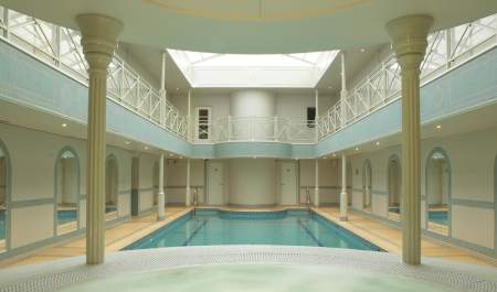 View from a hot tub of a rectangular pool surrounded by arched mirrors and ornamental pillars, with a mezzanine balcony above. The Lygon Arms Spa in the Cotswolds