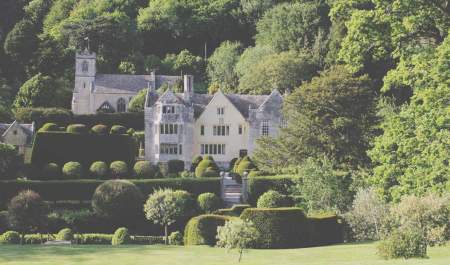 A Tudor manor house stands amidst formal gardens with a church perched higher up the valley in the distance at Owlpen Manor Cottages in the Cotswolds