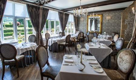 A bright dining room dressed in muted tones, chandeliers and mirrors, with large windows overlooking the gardens at The Greenway Hotel & Spa in the Cotswolds