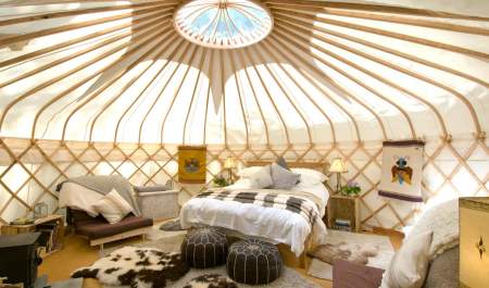 A circular skylight illuminates a cozy double bed, faux animal skin rugs and pouffes dotted in front of a wood burner inside Sapperton Yurt, at Westley Farm in the Cotswolds