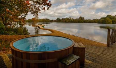 A wooden hot tub on a deck with views across the lake and leaves falling from the trees, at Log House Holidays in the Cotswolds