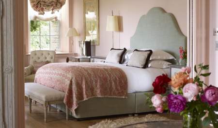 A vase of pink peonies sits on a table in front of a view of a large bedroom with upholstered bed, pale pink walls and large sash window at Thyme in the Cotswolds