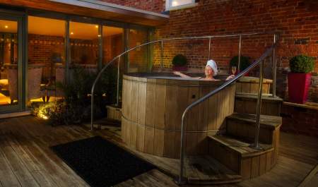 A lady relaxes at night in an outdoor wooden hot tub, surrounded by plants and subdued lighting at The Greenway Hotel & Spa in the Cotswolds