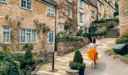 A woman wearing a hat smiles as she poses in front of stone cottages on a stepped hill in Tetbury