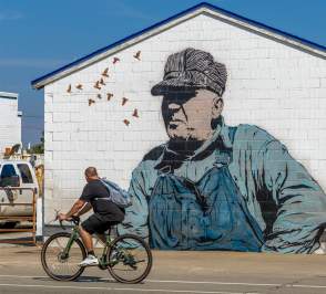 A man biking past a mural of an older working man titled Keep em Rollin by Chris Stain from the BOOM! Salina festival in Salina, Kansas