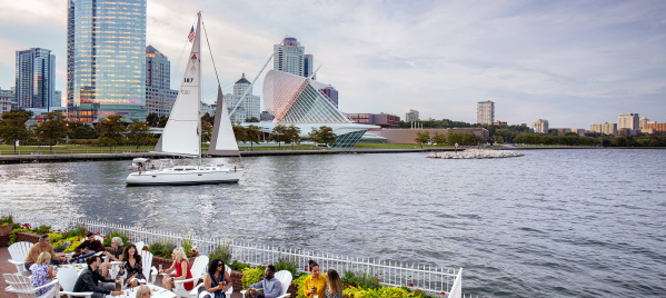 a group of people eating on the patio in front of the Milwaukee skyline with a sailboat in the harbor