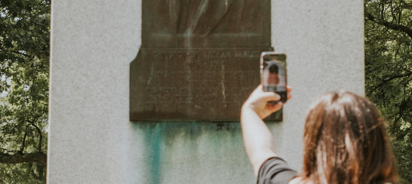 A person holds up a smartphone to take a photo of a stone monument with a bronze relief portrait plaque on it, surrounded by trees in the background.