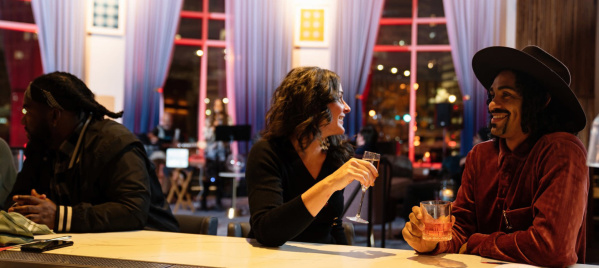 Two people sit at a stylish bar engaged in a lively conversation, each holding a drink. The woman smiles while talking to the man in a wide-brimmed hat. The setting is warmly lit with modern decor, including large windows, blue curtains, and ambient city lights in the background. Another person sits further down the bar, looking away.