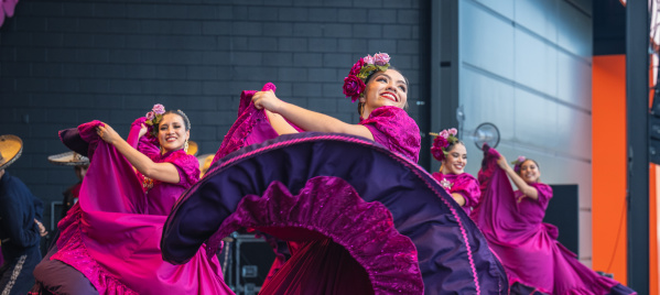 A group of female dancers performs a traditional Mexican folklórico dance on stage. They wear vibrant magenta dresses with purple ruffles and floral headpieces. The lead dancer in the foreground smiles brightly while gracefully twirling her flowing skirt. The dancers behind her mirror the movement, creating a colorful, energetic scene against a modern stage backdrop.
