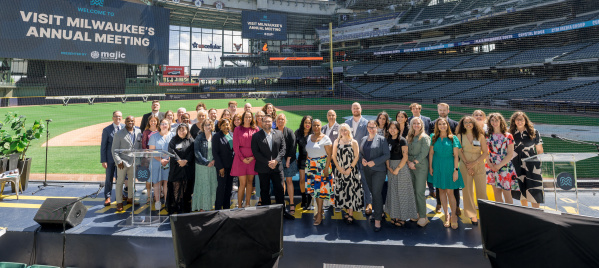 Group photo of attendees at Visit Milwaukee's Annual Meeting, posed on a stage at American Family Field (Milwaukee Brewers stadium) with stadium seating and banners visible in the background.