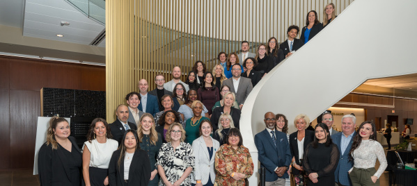 Large group of professionals posing on a curved staircase inside a modern building with wood slat walls and glass railings.