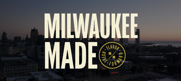 Aerial view of Milwaukee's downtown skyline at dusk with tall buildings silhouetted against a gradient sky. Large cream-colored text reads 'MILWAUKEE MADE' overlaid on the cityscape, with a circular yellow badge logo containing 'FRESH FLAVOR FORWARD' and stylized 'MM' letters positioned to the right of 'MADE'.