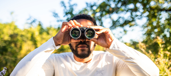 A man in a white long-sleeved shirt holds binoculars up to his eyes while standing outdoors in a sunny, tree-filled setting with green foliage and dappled sunlight in the background.