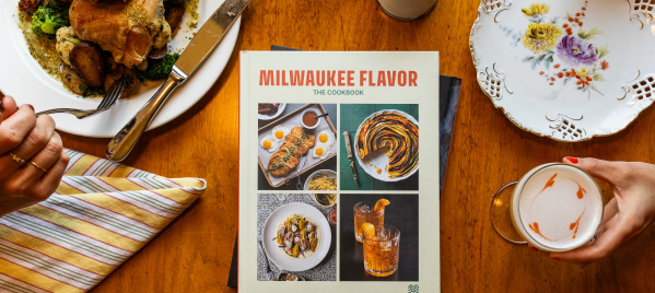 Milwaukee Flavor: The Cookbook rests on a wooden dining table surrounded by a meal. A hand holds a fork over a plate of chicken, vegetables, and potatoes on the left, while another hand holds a latte with heart-shaped foam art on the right. Around the book are a striped napkin, a floral plate, and a dessert plate with slices of cake.