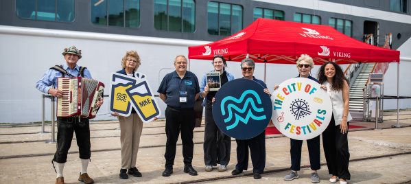 A welcoming committee with an accordion player stands before a cruise ship, holding Milwaukee-themed signs including an MKE logo and "City of Festivals" circular display, under a red Viking Cruises tent.