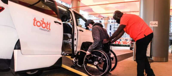 A man in a wheelchair uses a ramp to board a white accessible vehicle labeled "Tootl" while being assisted by a staff member in an orange shirt outside a brightly lit building.